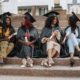 Group of young African female students dressed in black graduation gowns. Campus as a background. Girls sitting on a stairs and talking.