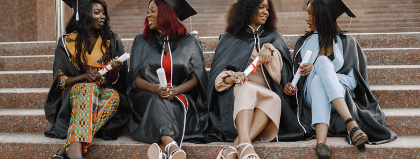 Group of young African female students dressed in black graduation gowns. Campus as a background. Girls sitting on a stairs and talking.