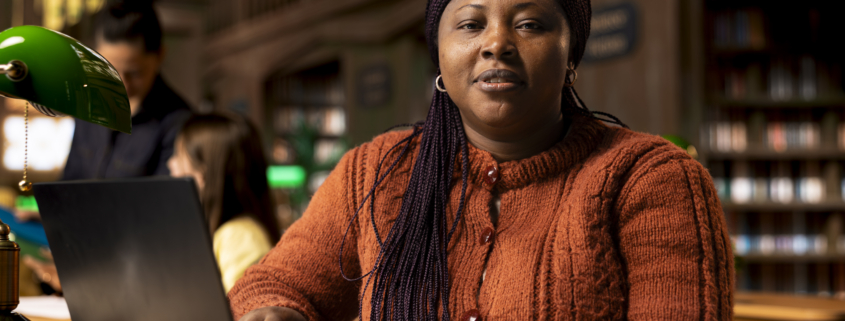Portrait of african teacher working with academic resources and learning materials within the public library database. Smiling woman focused on her educational profession.