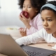 Two siblings, one eating an apple and the other one is on the laptop learning on an education portal