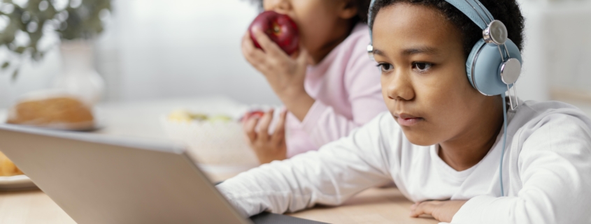 Two siblings, one eating an apple and the other one is on the laptop learning on an education portal