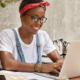 Stylish businesswoman works on laptop computer in cozy coffee shop, keyboards information, messages in social networks, has happy expression, wears glasses for good vision, focused into monitor