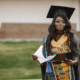 Young African female student dressed in a black graduation gown. Campus as a background. Girl holding some documents and looking at camera.