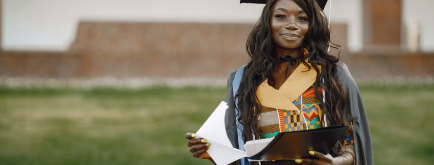 Young African female student dressed in a black graduation gown. Campus as a background. Girl holding some documents and looking at camera.