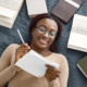 Young black woman lying down on bed writing down her notes at home. Girl wearing eyeglasses and beige sweater. Girl preparing her homework.