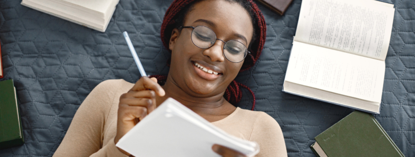 Young black woman lying down on bed writing down her notes at home. Girl wearing eyeglasses and beige sweater. Girl preparing her homework.